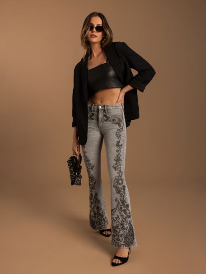 A model wearing light-wash flared jeans with black floral lace applique down the legs, styled with a black leather crop top, black blazer, black sunglasses, and black heels against a solid tan studio background.