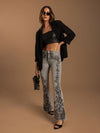 A model wearing light-wash flared jeans with black floral lace applique down the legs, styled with a black leather crop top, black blazer, black sunglasses, and black heels against a solid tan studio background.