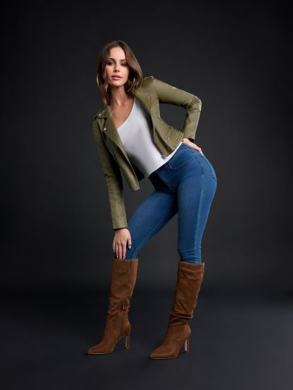 A woman poses in a studio wearing an olive green moto jacket, a white V-neck top, blue skinny jeans, and brown knee-high heeled boots against a dark gray background.