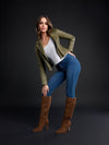 A woman poses in a studio wearing an olive green moto jacket, a white V-neck top, blue skinny jeans, and brown knee-high heeled boots against a dark gray background.