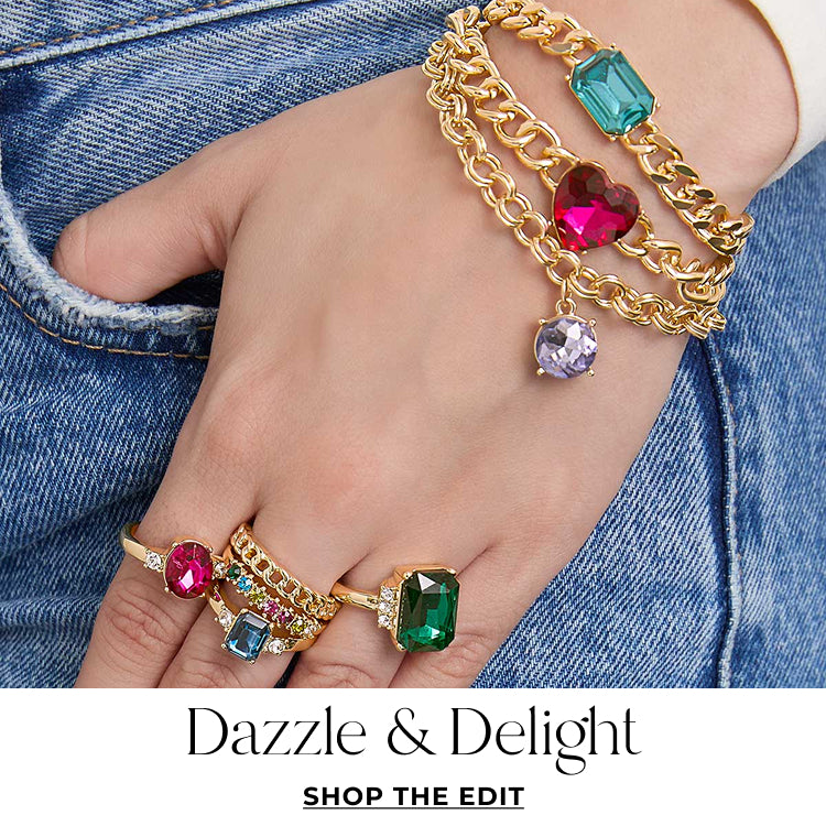 Gold jewelry with colorful gemstones on a hand against a denim background, featuring 'Dazzle & Delight' text.Shop the edit.
