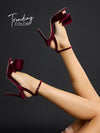 A pair of women's high-heeled sandals in deep burgundy velvet, featuring a large bow on the outer side and a thin ankle strap, shown on a model's feet against a dark gray background with 'Trending COLOR' text in the upper left.